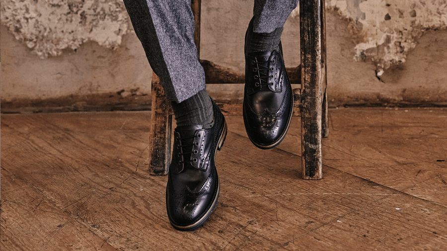 A man's feet resting on a wooden stool, wearing the LANX, Hayhurst // Black, hi-shine, derby brogues.