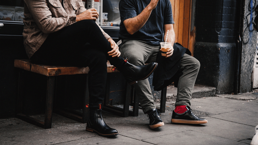Two men sat outside a coffee shop, drinking coffee. One wearing the LANX, Garstang // Black, hi-shine, chelsea boots and the other wearing the LANX, Ancoats // Gum & Black, hi-shine, leather sneakers with a gum sole.