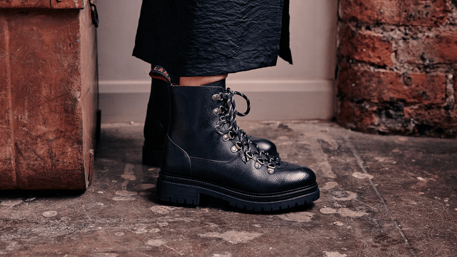 A woman wearing the LANX Whalley Black Grained, lace-to-toe boots. Stood on a conrete floor in a redbrick room.