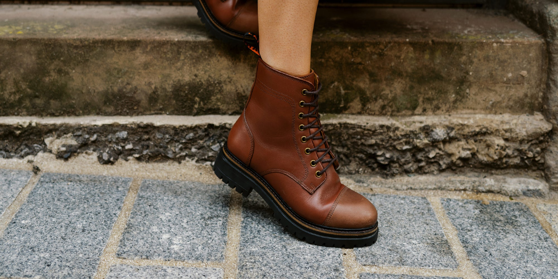 A woman wearing the LANX Whalley Black Grained, lace-to-toe boots. Stood on a conrete floor in a redbrick room.